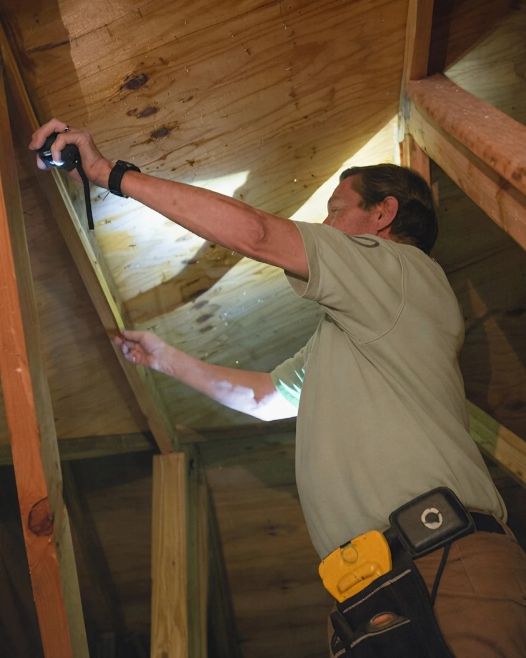 Roof inspection being performed by a man on the shingle roof of a single-story home, holding surveying tools, with an aluminum ladder leaning against the white stucco wall.